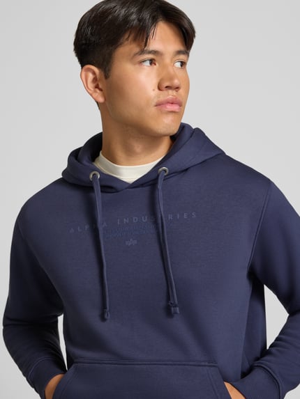 Mannelijk model draagt Relaxed Fit Hoodie uit katoenmix met labeldetail van Alpha Industries in marineblauw, Nahaufnahme Oberkörper. Heren hoodie, katoenmix, relaxed fit, kangoeroezak, capuchon met tunnelkoord, labeldetail, elastische boorden, ruglengte maat M 69 cm, mouwlengte maat M 62 cm.