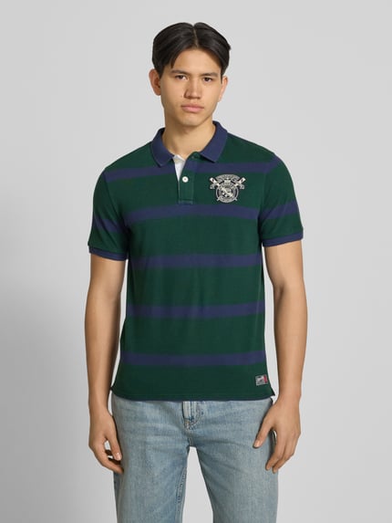 Männermodel trägt Slim Fit Poloshirt mit Label-Stitching und Knopfleiste von Superdry aus Reine Baumwolle, grün-blau gestreift, Slim-Fit Herren, Rückenlänge bei Größe M 70 cm, Ganzkörperansicht von vorne.