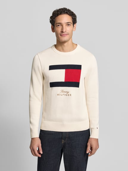 Männermodel trägt Regular Fit Pullover mit Motiv-Print von Tommy Hilfiger in Strickoptik, Rundhals, langarm, Logo-Detail, Ganzkörperansicht von vorne.