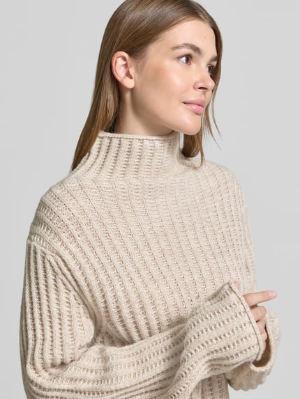 Frauenmodel trägt Strickpullover mit Stehkragen Modell 'ARWIE' von DRYKORN, beiger Material-Mix mit Alpaka-Anteil, Nahaufnahme Oberkörper.