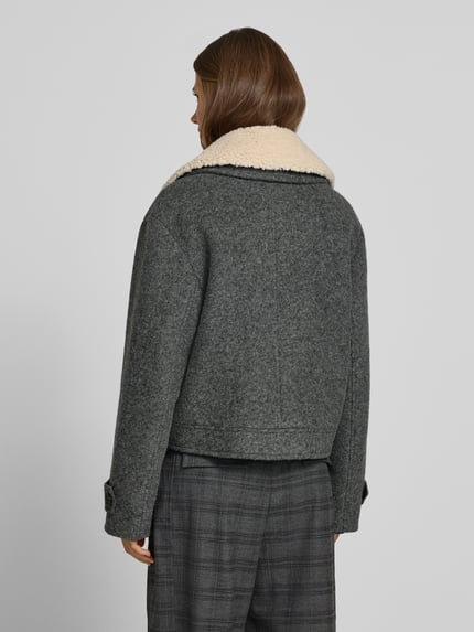 Frauenmodel trägt Jacke mit Kentkragen und Eingrifftaschen von Style Icon Collection, graue Farbe, strukturierte Oberfläche, Ganzkörperansicht Rückansicht. Gerader Schnitt, Knopfleiste, seitliche Eingrifftaschen, Ärmelriegel mit Knopf, Rückenlänge 54 cm, Schulterbreite 52 cm, Brustweite 50 cm, Ärmellänge 60 cm.