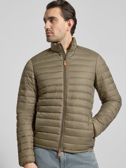 Männermodel trägt olivgrüne Steppjacke mit Zweiwegereißverschluss Modell 'Alexander' von SAVE THE DUCK, Stepp-Optik, Regular Fit, gefütterter Stehkragen, Eingrifftaschen mit Reißverschluss, Ganzkörperansicht von vorne.