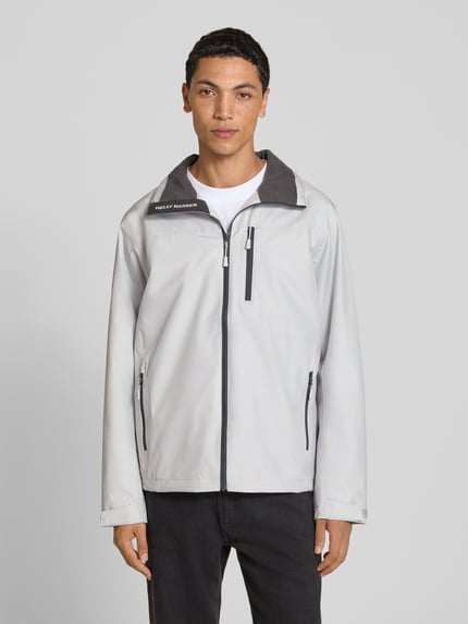 Männermodel trägt Regular Fit Jacke mit Stehkragen Modell 'Crew' von Helly Hansen, hellgrau mit schwarzem Reißverschluss, Ganzkörperansicht von vorne. Kontext: Herren Jacke, Stehkragen, Regular Fit, Modell 'Crew', Logo-Print, Napoleontasche, Eingrifftaschen mit Reißverschluss, Rückenlänge bei Größe M 73 cm, Schulterbreite 48 cm, Brustweite 56 cm, Ärmellänge 68 cm.
