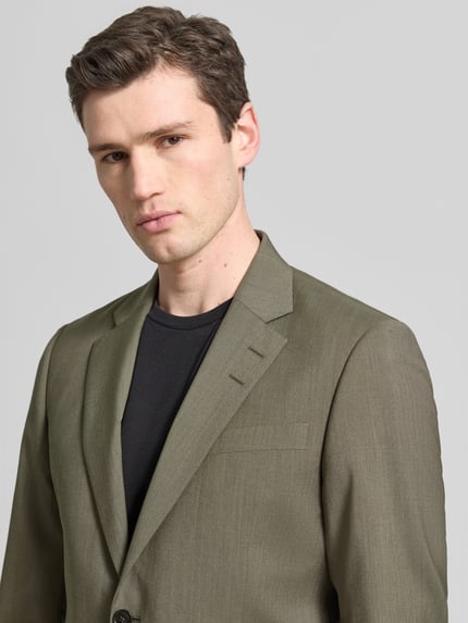 Männermodel trägt Regular Fit Blazer mit Logo-Patch Modell 'Justin' van Tiger Of Sweden in olijfgroen, puur katoen, borstzak, reverskraag, 2-knoopsluiting, detailaufnahme bovenlichaam, context heren blazer, ruglengte maat 50: 79 cm, schouderbreedte: 38 cm, mouwlengte: 66 cm.