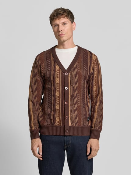 Męski model nosi Relaxed Fit Strickjacke aus reiner Baumwolle mit Knopfleiste od COLUCCI, bawełna, krój relaxed fit, listwa guzikowa, dekolt w serek, detal z logo, długość tyłu w rozmiarze M: 67 cm, długość rękawów w rozmiarze M: 67 cm, brązowe odcienie, geometryczne wzory, tekstura dzianiny, całkowity widok z przodu.