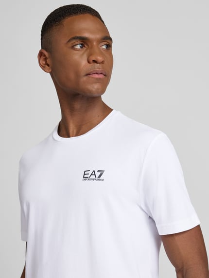 Männermodel trägt T-Shirt mit Label-Print von EA7 aus Baumwoll-Elasthan-Mix, Slim-Fit, Weiß, gerippter Rundhalsausschnitt, Nahaufnahme Oberkörper.