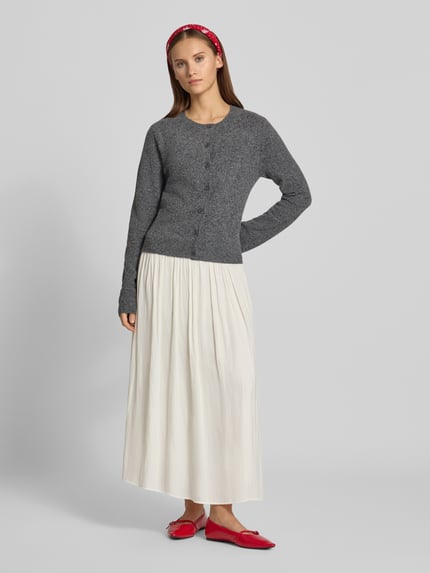 Frauenmodel trägt Regular Fit Strickjacke mit Lammwoll-Anteil von REVIEW, grau, Knopfleiste, Langarm, gerippte Abschlüsse, weißer Rock, Ganzkörperansicht von vorne.