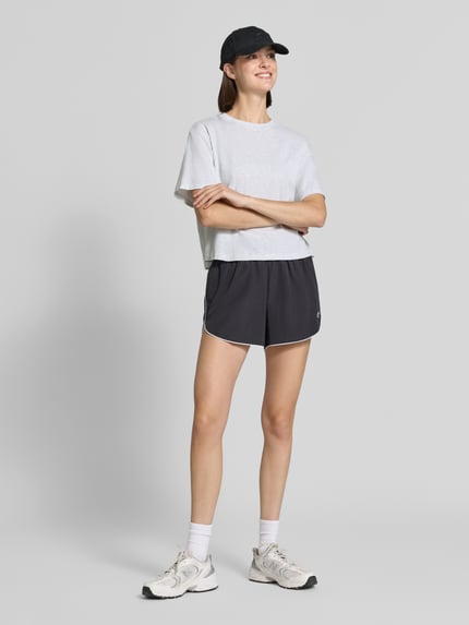 Frauenmodel trägt Relaxed Fit T-Shirt aus Baumwoll-Mix von Fabletics in Hellgrau, kombiniert mit Shorts, Ganzkörperansicht von vorne. Elastischer Baumwoll-Mix, Rundhalsausschnitt, überschnittene Schultern, kurzarm, Rückenlänge bei Größe S 48 cm.