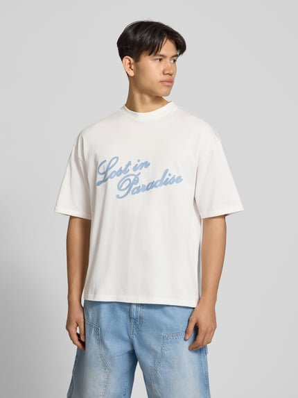 Männermodel trägt T-Shirt mit Logo- und Statement-Stitching von REVIEW aus reiner Baumwolle, gerader Schnitt, helle Farben, Ganzkörperansicht von vorne.