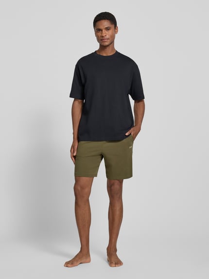 Männermodel trägt Regular Fit Sweatshorts aus Baumwoll-Mix Modell 'MIX&MATCH' von BOSS in Oliv, kombiniert mit schwarzem Shirt, Herren Sweatshorts, Ganzkörperansicht von vorne.