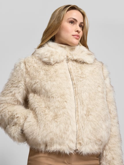 Modelka kobieca nosi Jacke aus Kunstfell mit Reißverschluss od Stefanel w kolorze jasnego beżu, regular fit, kołnierz wykładany, zamek błyskawiczny, tekstura miękkiego sztucznego futra; widok na górną część ciała z przodu.