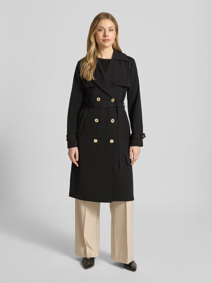 Modelka nosi czarny dwurzędowy trencz ‘Zweireihiger Trenchcoat mit Bindegürtel’ od MICHAEL Michael Kors, krój regular fit, kołnierz z połami, wiązany pasek, długość tyłu w rozmiarze S: 47 cm, szerokość ramion w rozmiarze S: 40 cm, szerokość klatki piersiowej w rozmiarze S: 50 cm, długość rękawów w rozmiarze S: 63 cm, jasne spodnie, prosty krój, gładka faktura, widok całej sylwetki z przodu.