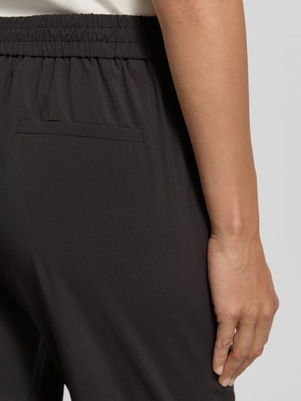 Vrouwenmodel draagt Hose in verkürzter Passform Modell 'Charlie' van Someday in zwart materiaalmix met stretch, elastische band, achterzak, detailopname heup. Context: dames broek met verkorte pasvorm, binnenbeenlengte 67 cm, bandwijdte maat 36 is 39 cm.