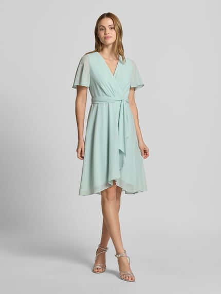 Frauenmodel trägt Knielanges Cocktailkleid in Wickel-Optik von Christian Berg Cocktail aus mintfarbenem Chiffon mit A-Linie, V-Ausschnitt, Bindegürtel, knielang, Ganzkörperansicht von vorne.