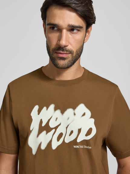 Männermodel trägt T-Shirt mit Label-Print Modell 'Bobby' von Wood Wood in Braun mit weißem Print, reine Baumwolle, Nahaufnahme Oberkörper.