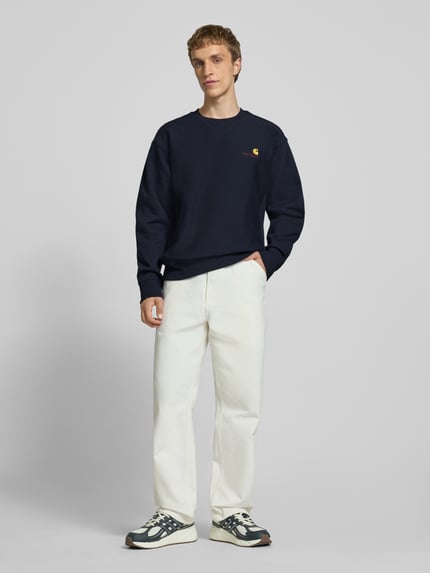 Männermodel trägt Regular Fit Sweatshirt mit Logo-Stitching von Carhartt Work In Progress in Dunkelblau, Baumwoll-Mix, Rundhalsausschnitt, weiße Hose, Ganzkörperansicht von vorne.