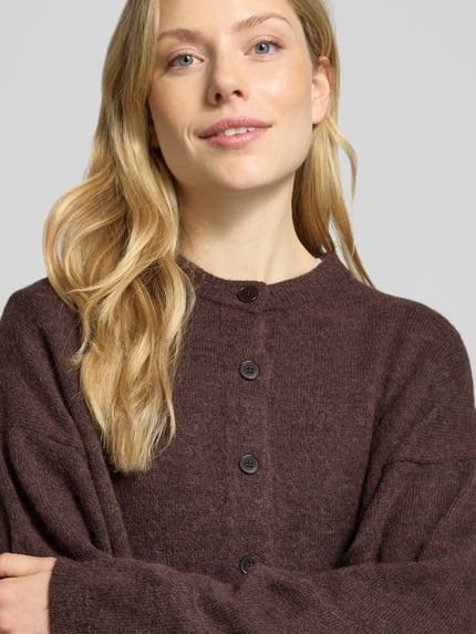 Frauenmodel trägt Cardigan aus Alpaka-Mix Modell 'MARWENI' von DRYKORN, braune Farbe, strukturierte Oberfläche, Nahaufnahme Oberkörper. Relaxed Fit, Rundhalsausschnitt, Knopfleiste, Rückenlänge 51 cm, Ärmellänge 70 cm, Woll-Anteil.