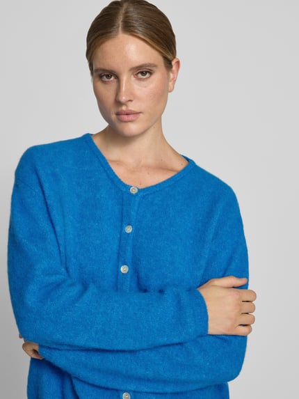 Frauenmodel trägt Relaxed Fit Strickjacke aus Woll-Mix mit Leinen-Anteil von American Vintage in leuchtendem Blau, gerippte Bündchen, durchgehende Knopfleiste, langärmlig, Nahaufnahme Oberkörper.