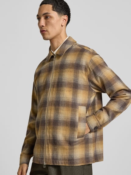 Männermodel trägt Regular Fit Overshirt aus Woll-Mix von Forét, kariert in Beige, Braun und Grau, Umlegekragen, Eingrifftaschen, Nahaufnahme Oberkörper.