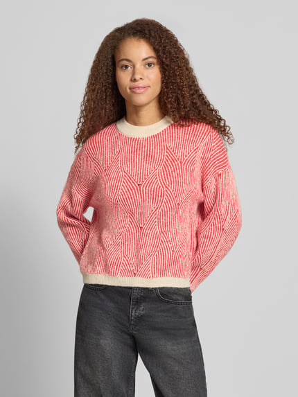 Frauenmodel trägt Pullover mit strukturiertem Jacquard-Strick von PIECES in Rot und Creme, geometrisches Allover-Muster, Strickoptik, Rundhalsausschnitt, lange Ärmel, gerippte Abschlüsse, Ganzkörperansicht von vorne.