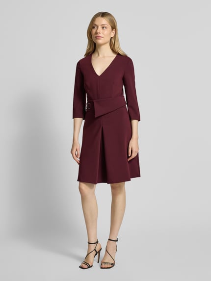 Frauenmodel trägt Knielanges Cocktailkleid mit V-Ausschnitt von Rinascimento in Weinrot, Material-Mix mit Viskose-Anteil, Taillierter Schnitt, Knielänge, V-Ausschnitt, Taillengürtel, 3/4-Ärmel, Nahtreißverschluss, Ganzkörperansicht von vorne.