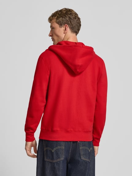 Männermodel trägt Regular Fit Sweatjacke aus Baumwoll-Mix mit Label-Detail von Superdry in Rot, Rückansicht, Kontext: Kapuze, Känguru-Tasche, elastische Abschlüsse, Rückenlänge 65 cm, Ärmellänge 65 cm.