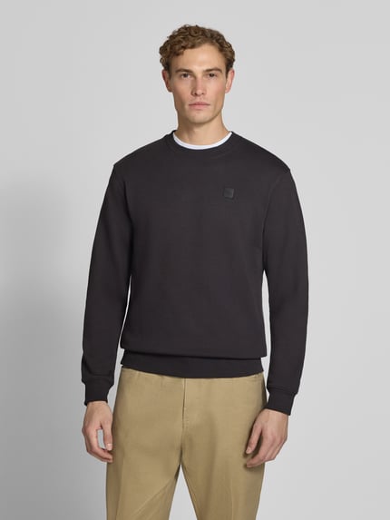 Męski model nosi Regular Fit Sweatshirt aus reiner Baumwolle mit Label-Patch od Scotch & Soda, krój regular fit, czarna bawełna, elastyczne wykończenia, naszywka z logo, długość tyłu w rozmiarze M 70 cm, długość rękawów 66 cm, zdjęcie całej sylwetki z przodu.