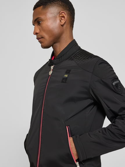 Męski model nosi Jacke mit Zweiwege-Reißverschluss Modell 'Thayer' od Blauer Usa, czarny materiał stretch, widoczne kieszenie, naszywka na rękawie, zbliżenie na górną część ciała, ujęcie z przodu. Szerokość w rozmiarze M: 54 cm.