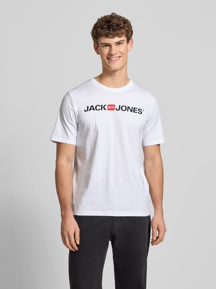 Męski model nosi T-Shirt z nadrukiem label-Print model 'CORP' od JACK & JONES, biały kolor, prosty krój, bawełna z wiskozą, ujęcie całej sylwetki z przodu.