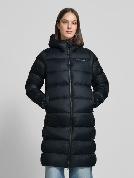 Frauenmodel trägt schwarzen Steppmantel mit Daunen-Federn-Füllung Modell 'FROST' von Peak Performance, Regular Fit, Kapuze, Eingrifftaschen, Label-Stitching, Reißverschluss, Ganzkörperansicht von vorne.