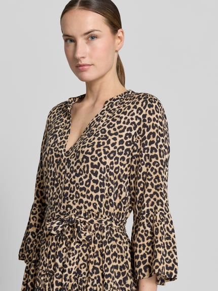Frauenmodel trägt tailliertes Tunikakleid aus reiner Viskose Modell 'MELBA' von Vero Moda mit Allover-Leo-Muster, Trompetenärmeln, Volantsaum, Bindegürtel, Mini-Länge; Nahaufnahme Oberkörper.