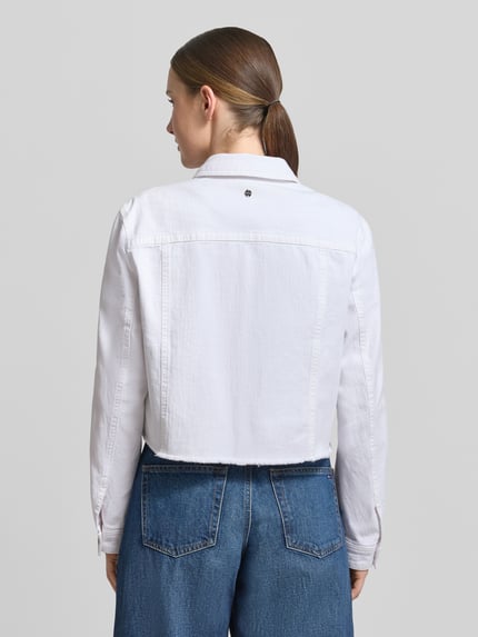 Frauenmodel trägt Jeansjacke mit Brusttasche und Fransen von Marc Cain aus weißem Baumwoll-Elasthan-Mix, Ganzkörperansicht Rückansicht, Slim-Fit, Umlegekragen, Knopfleiste, Fransen, Rückenlänge bei Größe 36: 47 cm, Schulterbreite 39 cm, Brustweite 37 cm, Ärmellänge 59 cm.