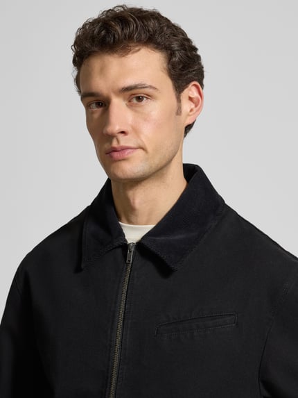 Männermodel trägt Jacke mit Zweiwege-Reißverschluss Modell 'Bo' von Wood Wood aus schwarzer Baumwolle, Nahaufnahme Oberkörper. Regular Fit, Umlegekragen, sichtbarer Reißverschluss und Brusttasche.