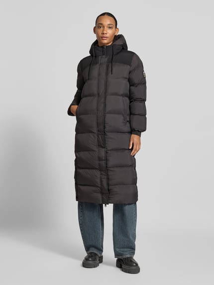 Frauenmodel trägt Steppmantel mit Kapuze und verdecktem Reißverschluss von Superdry in Schwarz, Daunenoptik, gerade Form, Eingrifftaschen, Ganzkörperansicht von vorne. Rückenlänge 108 cm, Schulterbreite 42 cm, Brustweite 53 cm, Ärmellänge 61 cm, Regular Fit.