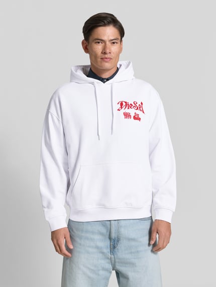 Männermodel trägt Oversized Hoodie aus reiner Baumwolle von Diesel in Weiß mit rotem Label-Print, Känguru-Tasche, Ganzkörperansicht von vorne.