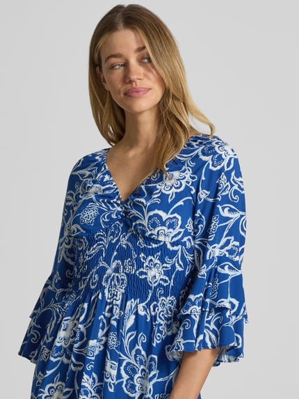 Frauenmodel trägt Minikleid aus reiner Viskose mit Smok-Detail von Apricot, floraler Allover-Print in Blau und Weiß, Rüschenbesatz, 3/4-Arm, V-Ausschnitt, taillierte Passform, Nahaufnahme Oberkörper.