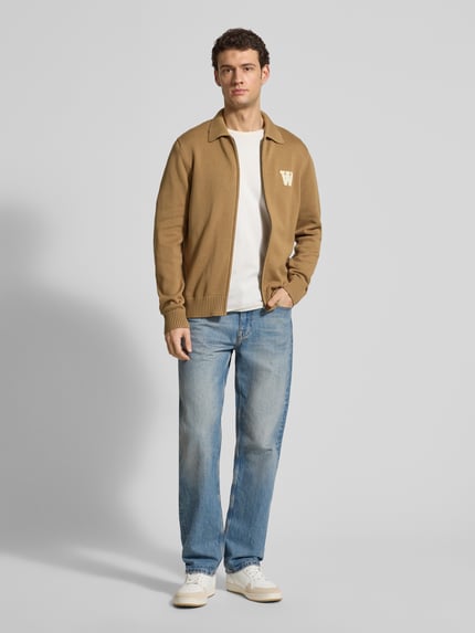 Männermodel trägt Regular Fit Cardigan in Strick-Optik Modell 'Tay' von Wood Wood, beige, Umlegekragen, Reißverschluss, Ganzkörperansicht von vorne.