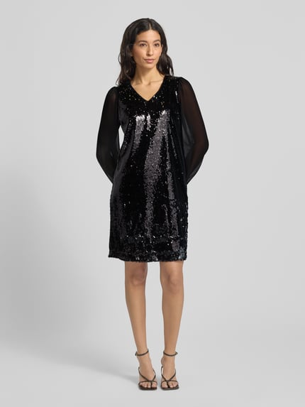 Kobiece modelka nosi Knielanges Kleid mit Paillettenbesatz von More & More, czarny połysk, elastyczna mieszanka materiałów, taliowany krój, długość do kolan, dekolt w serek, wycięcie w kształcie łezki z tyłu, zapięcie na guzik, półprzezroczyste bufiaste rękawy, przód z cekinami, widok całej sylwetki od przodu.