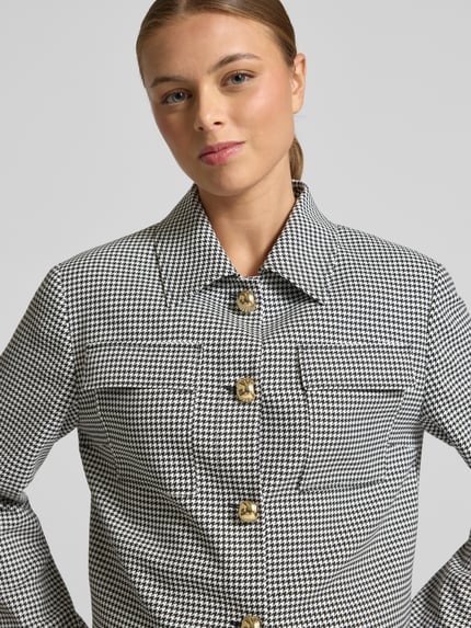 Vrouwenmodel draagt Boxy Fit Kurzblazer met Vichy-muster Modell 'ALERO-2' van HUGO in zwart-wit, allover patroon, borstpattentas. Nahaufnahme bovenlichaam.