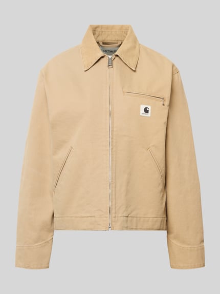 Produktfoto, Jacke mit Label-Detail von Carhartt Work In Progress aus beigefarbener Baumwolle, entspannter Schnitt, sichtbare Reißverschlüsse, Umlegekragen, schlichte Struktur.