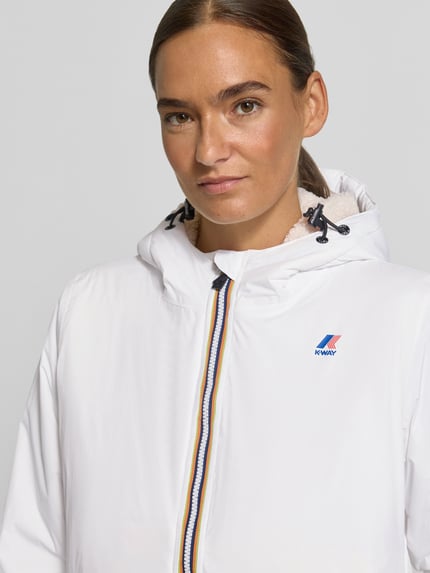 Frauenmodel trägt weiße Regular Fit Jacke mit Logo-Print Modell 'CLAUDETTE' von K-WAY, sichtbare Kapuze, Nahaufnahme Oberkörper. Damen Jacke, elastische Abschlüsse, Tunnelzug, glatte Textur.
