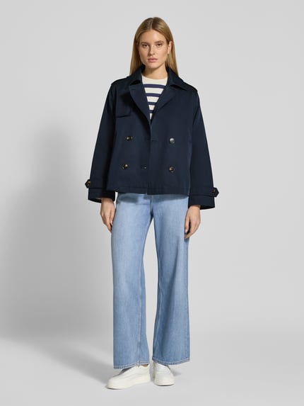 Modelka nosi Trenchcoat in kurzem Design mit Baumwoll-Anteil von Jake*s Casual, granatowy kolor, prosta forma, bawełniany materiał, kołnierz, podwójny rząd guzików, kieszenie, pagony na ramionach i rękawach, krótki fason, długość tyłu w rozmiarze 36: 64 cm, szerokość ramion w rozmiarze 36: 44 cm, szerokość klatki piersiowej w rozmiarze 36: 51 cm, długość rękawa w rozmiarze 36: 60 cm, widok całej sylwetki z przodu.