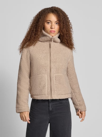 Frauenmodel trägt Teddyjacke mit hohem Stehkragen von ONLY aus beigem Fleece, unifarben, Strukturmuster, aufgesetzte Taschen, Ganzkörperansicht von vorne.