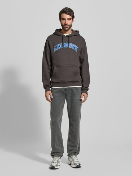 Männermodel trägt Hoodie mit Label-Print Modell 'Brady' von Les Deux aus reiner Baumwolle, Regular-Fit, Känguru-Tasche, Ganzkörperansicht von vorne.