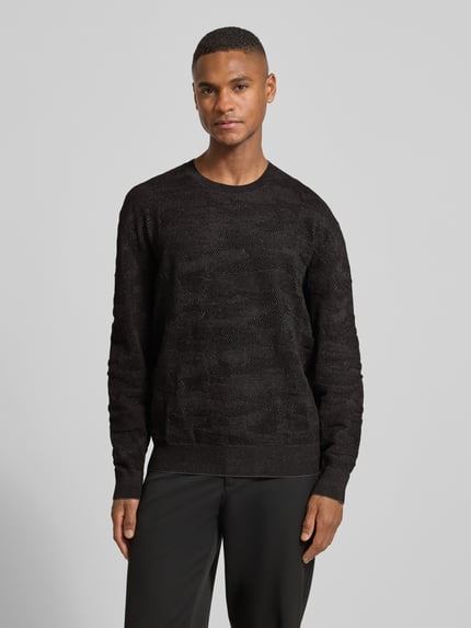 Männermodel trägt Strickpullover mit gerippten Abschlüssen von ARMANI EXCHANGE im schwarzen Material-Mix, lockerer Schnitt, Ganzkörperansicht von vorne.