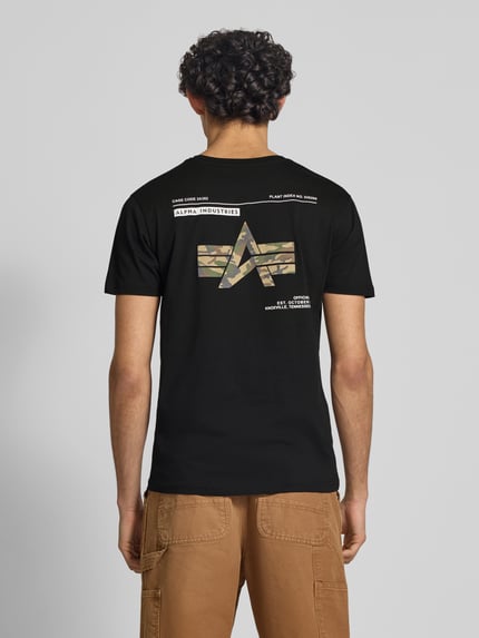 Mężczyzna model nosi T-Shirt mit Label-Patch Modell 'Camo' od Alpha Industries, czarna bawełna, prosty krój, detal z logo, widok z tyłu.