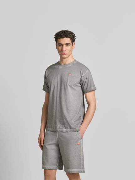 Męski model nosi Regular Fit T-Shirt aus reiner Baumwolle von Diesel, szary kolor, krój regular fit, okrągły dekolt, używany wygląd, detale z logo, długość pleców 70 cm w rozmiarze M, zdjęcie w całej sylwetce z przodu.