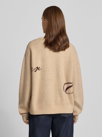 Frauenmodel trägt Oversized Strickpullover mit Label-Details von ANOTHER COTTON LAB in Beige mit Rundhalsausschnitt, strukturierte grobe Textur, Rückenansicht, Kontext: Damen Strickpullover, überschnittene Schultern, Rückenlänge 60 cm, Ärmellänge 62 cm.