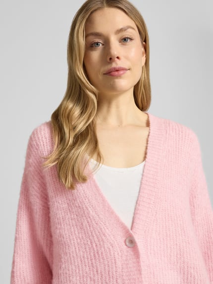 Frauenmodel trägt Cardigan mit V-Ausschnitt Modell 'EAST' von American Vintage in Hellrosa, grobe Strickstruktur, Nahaufnahme Oberkörper. Relaxed Fit, Knopfleiste, Rückenlänge 62 cm, Ärmellänge 48 cm bei XS/S.