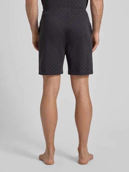 Mężczyznęmodel nosi czarne szorty aus Baumwolle mit Logo-Muster von JOOP! Collection, krój regular fit, wzór z logo, elastyczny pas, szerokość w rozmiarze M: 42 cm, długość wewnętrzna nogawki: 18 cm, widok całej sylwetki z tyłu.
